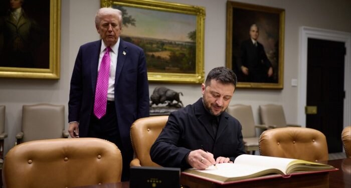 Volodymyr Zelensky signs a book at a table while the US president stands behind him