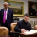 Volodymyr Zelensky signs a book at a table while the US president stands behind him