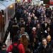 Commuters crowd a platform at Waterloo Station in London during rush hour, with a train on the left and people walking in both directions.