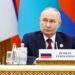 Vladimir Putin sits at a table with a sign reading "Russian Federation" during an international forum, with colorful flags in the background.