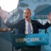 Nigel Farage stands at a podium with arms raised, speaking passionately at a Reform UK rally in front of a blue campaign backdrop.
